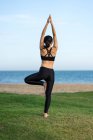 Visão traseira da mulher nova no alto preto e os leggings que estão na pose da árvore na grama verde ao praticar o yoga na praia — Fotografia de Stock