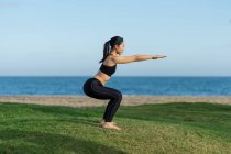 Giovane donna in top nero e leggings in piedi su erba verde praticare yoga sulla spiaggia — Foto stock