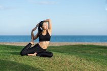 Young woman in black top and leggings sitting on green grass practicing yoga on beach — Stock Photo