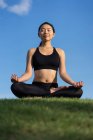 Young woman in black top and leggings sitting on lotus position on green grass with closed eyes while meditating at the beach — Stock Photo