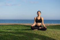 Woman in black top and leggings sitting on lotus position on green grass with closed eyes while meditating at the beach — Stock Photo