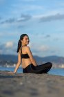 Side view of woman in black sportswear sitting with crossed legs on beach resting after workout looking at camera — Stock Photo