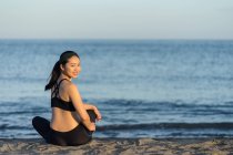 Вид збоку усміхненої жінки в чорному спортивному одязі, що сидить з схрещеними ногами на пляжі, відпочиваючи після тренування, дивлячись на камеру — стокове фото