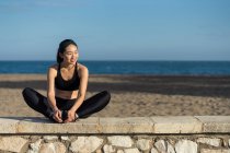 Молодая азиатская женщина в черном топе и с ногами, делающая упражнения на растяжку, сидя рядом с каменным забором в стороне — стоковое фото