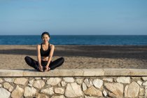 Jovem asiático mulher no preto topo e leggings fazendo alongamento exercício enquanto sentado ao lado de cerca de pedra à beira-mar — Fotografia de Stock