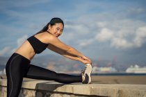 Side view of Asian woman in black top and leggings doing stretching exercise while standing next to stone fence at seaside — Stock Photo