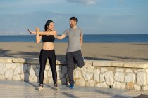 Pareja de pie en una de las piernas mientras se mantienen unos a otros en ropa deportiva con mar azul y cielo sobre fondo borroso. - foto de stock