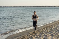 Atleta joven motivada con traje negro activo y zapatillas que juegan a lo largo de la arena de mar vacía. - foto de stock