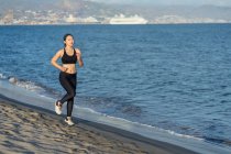Atleta feminina motivada em desgaste preto ativo e tênis correndo ao longo de praia vazia arenosa — Fotografia de Stock