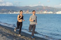 Couple en vêtements de sport souriant tout en joggant sur le côté sablonneux de l'océan avec la ville sur la côte opposée sur fond flou — Photo de stock