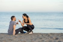 Alegre jovem casal multirracial no sportswear sentado na praia de areia, enquanto descansa após o treinamento e desfrutar de tempo juntos — Fotografia de Stock