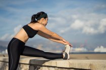 Side view of young Asian female in black top and leggings doing stretching exercise while standing next to stone fence — Stock Photo