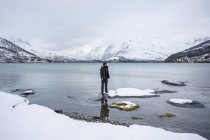 Solitary tourist on fjord shore against snowy hills in cold overcast weather — Stock Photo