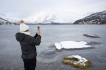 Frau schießt bei kaltem Wetter mit Smartphone am Teichstrand gegen verschneite Hügel — Stockfoto