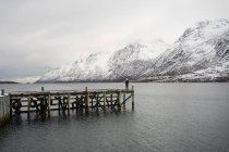 Lonely tourist standing on wooden pier in middle of calm lake in winter — Stock Photo