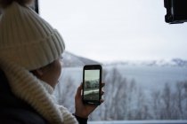 Calm woman recording video with smartphone of wintry scenery while looking out window — Stock Photo