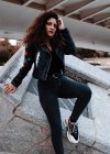 Mujer moderna en chaqueta negra y jeans de pie en la ciudad - foto de stock