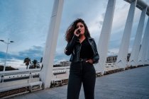 Стильная женщина в черной куртке и джинсах курит на мосту — стоковое фото