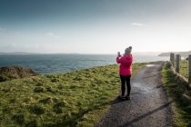 Mujer de pie en la carretera del país y tomar fotos del mar con el teléfono inteligente - foto de stock