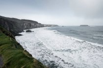 Paysage marin à couper le souffle du littoral d'Irlande du Nord avec des roches et de l'herbe verte de printemps et des vagues froides orageuses de l'océan se brisant sur le rivage avec de la mousse — Photo de stock