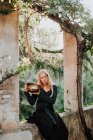 Mulher de vestido longo roupas com olhos fechados e segurando tigela musical de metal enquanto sentado em ruínas do castelo velho no lugar histórico — Fotografia de Stock
