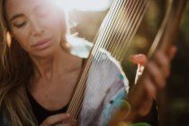 Романтическая очаровательная женщина с светлыми волосами, наслаждающаяся мелодией, играя на музыкальном инструменте и сидя в саду летом — стоковое фото