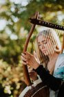 Mujer encantadora romántica con cabello rubio disfrutando de la melodía mientras toca el instrumento musical y se sienta en el jardín en verano - foto de stock