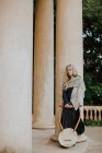 Женщина-музыкант в повседневной одежде отдыхает после выступления и держит деревянную лиру, опираясь на колонну у входа в здание — стоковое фото