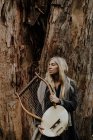 Tender woman with blond hair holding wooden antique musical instrument while standing with closed eyes at old tree trunk — Stock Photo