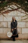Pensativo encantador loiro músico feminino no casaco segurando instrumento musical corda enquanto em pé na parede de pedra velha no outono — Fotografia de Stock