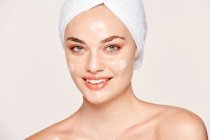 Donna lentigginosa positiva con asciugamano sulla testa e pelle sana prendersi cura del viso con crema e guardando la fotocamera isolata su sfondo bianco — Foto stock