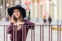 Content long haired woman in fashionable black hat and shirt leaning on fence while calling on mobile phone and looking at camera — Stock Photo