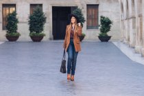 Donna eccitata in elegante abbigliamento casual e cappello nero a piedi e guardando lontano sul marciapiede tra vecchi edifici — Foto stock