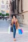 Satisfied woman in stylish casual outfit and hat walking with sopping bags and looking away at city street — Stock Photo