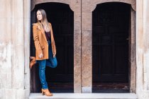 Satisfied charming woman in stylish casual clothing and heels leaning on wall of building with arch and looking away at city street — Stock Photo