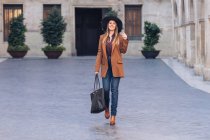 Mulher excitada em elegante desgaste casual e chapéu preto andando e olhando para a câmera na calçada entre edifícios antigos — Fotografia de Stock