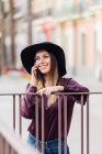 Zufriedene langhaarige Frau mit modischem schwarzen Hut und Hemd lehnt am Zaun, während sie mit dem Handy telefoniert und wegschaut — Stockfoto