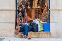Stylische langhaarige Frau mit Hut Shopaholic in lässiger modischer Kleidung sitzt mit Einkaufstaschen auf geschaukelten Zaun — Stockfoto