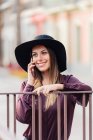 Вміст довгошерстої жінки в модному чорному капелюсі і сорочці, спираючись на паркан під час виклику на мобільний телефон і озираючись — стокове фото