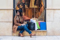 Стильная длинноволосая женщина в шляпе шопоголика в повседневной модной одежде сидит с сумками для покупок — стоковое фото