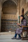 Вид сбоку на стильную женщину, сидящую на стуле и отдыхающую внутри старого исламского дворца с помощью мобильного телефона — стоковое фото