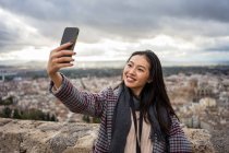 Mulher asiática feliz na roupa elegante tocar cabelo e tomar selfie enquanto está em pé contra a cidade envelhecida e céu nublado — Fotografia de Stock