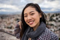 Щаслива азіатська жінка в стильному пальто і шарф посміхається і дивиться на камеру, стоячи на розмитому тлі старого міста — стокове фото