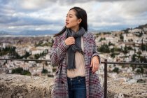 Счастливая женщина в стильном пальто и шарфе улыбается и смотрит в сторону, стоя на размытом фоне старого города — стоковое фото