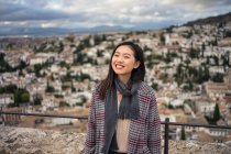 Счастливая женщина в стильном пальто и шарфе улыбается и смотрит в камеру, стоя на размытом фоне старого города — стоковое фото