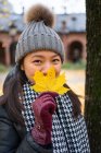 Asian woman in warm clothes looking at camera and covering face with yellow maple leaf while standing at Oslo Cathedral in Norway — Stock Photo
