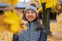 Una joven y alegre mujer asiática tirando hojas de arce amarillo mientras se encuentra en el patio de la catedral de Oslo, en Noruega, mirando hacia otro lado. - foto de stock