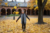 Una mujer asiática alegre tirando hojas de arce amarillo mientras se encuentra en el patio de la catedral de Oslo, en Noruega. - foto de stock