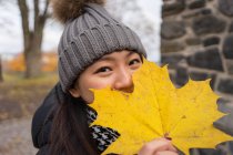 Mulher asiática em roupas quentes olhando para a câmera e cobrindo o rosto com folha de bordo amarelo enquanto está em pé na Catedral de Oslo, na Noruega — Fotografia de Stock