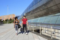 Donna afroamericana e uomo caucasico con borsa sportiva in mano camminando insieme lungo la parete della costruzione della città — Foto stock